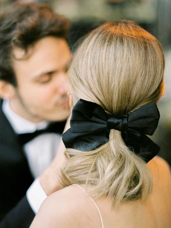 A close-up portrait of a couple sharing a kiss, shot from behind the bride to highlight her bridal hairstyle — a low, loosely gathered ponytail secured with a large black satin bow. The groom, visible in soft focus in the background, wears a black tuxedo with a black bow tie. The bride wears a thin-strap white dress. The styling is classic black-and-white formal with a graphic hair accessory as a focal point. The composition is a tight detail-portrait hybrid with intentional shallow depth of field.