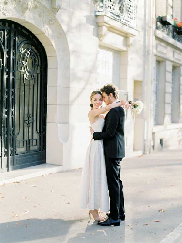 A couple portrait taken outdoors on a Parisian sidewalk in front of a classic Haussmann-style building with ornate wrought-iron doors and stone facade. The bride wears a tea-length ivory strapless dress with a textured skirt and nude heels, holding a small white bouquet, while the groom wears a black suit with a white dress shirt and no tie. The two are embracing closely, with the groom nuzzling into the bride's neck as she smiles toward the camera. The shot is a medium full-body portrait with warm, golden natural light casting soft shadows across the scene.