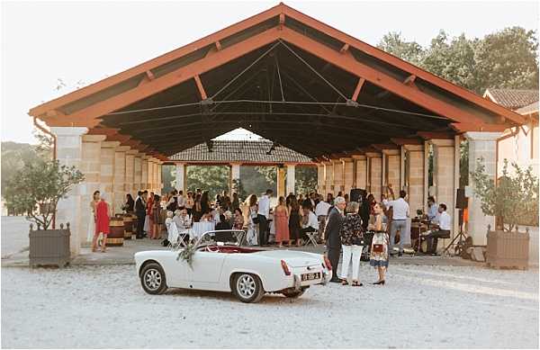 A cocktail hour is underway at an outdoor covered pavilion with a pitched wooden roof supported by stone columns, likely at a French chateau or domaine. Approximately 40–50 guests mingle beneath the open-sided structure, dressed in summer attire including bright and neutral tones. In the foreground, a vintage white convertible sports car with a red interior and a small white floral arrangement on its hood is parked on a gravel surface, serving as a decorative detail. The wide shot captures the warm golden evening light, a live band visible to the right side of the pavilion, and the overall relaxed, summery atmosphere of the reception. Potential venue feature image.