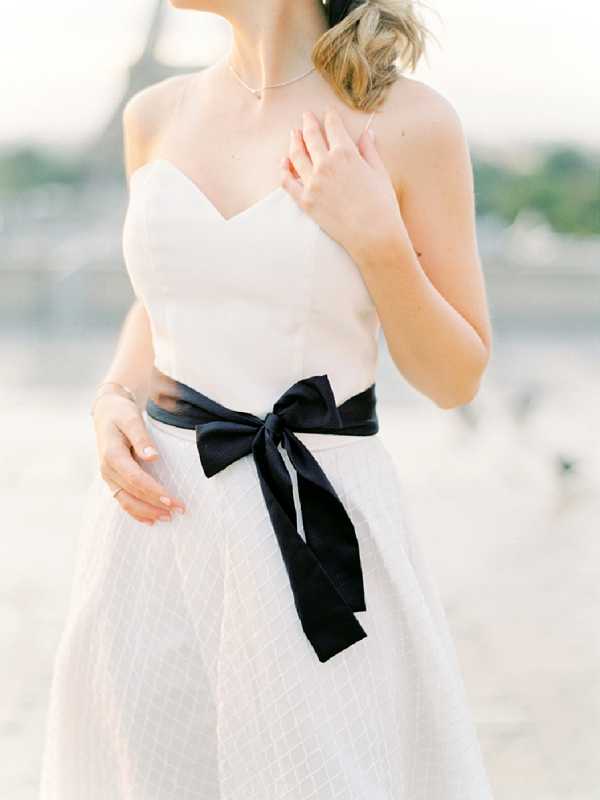A close-up portrait of a bride from the chin to mid-thigh, shot outdoors with a softly blurred background that suggests a Parisian setting, with what appears to be the Eiffel Tower faintly visible in the background. She wears a strapless white wedding dress with a sweetheart neckline and a textured quilted skirt, accessorized with a wide black satin sash tied in a large bow at the waist. A delicate silver chain necklace is visible at her collarbone, and she wears a ring on her right hand. The styling is clean and modern with a strong monochrome contrast between the white gown and the black ribbon detail.