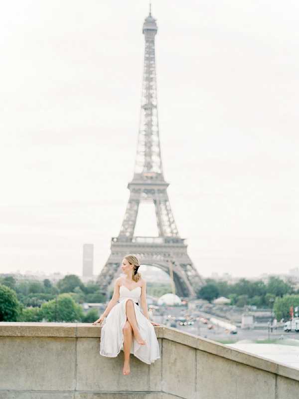 A bride sits alone on a stone ledge at the Trocadéro in Paris, with the Eiffel Tower centered directly behind her in soft focus. She is wearing a white strapless sweetheart-neckline gown with a flowing skirt featuring a front slit, and her hair is loosely styled in a low updo. The shot is a full-length portrait taken at a slight distance, with the bride positioned in the lower-left third of the frame to allow the tower to dominate the background. The overall image has a soft, muted, film-like quality with pale, diffused light.