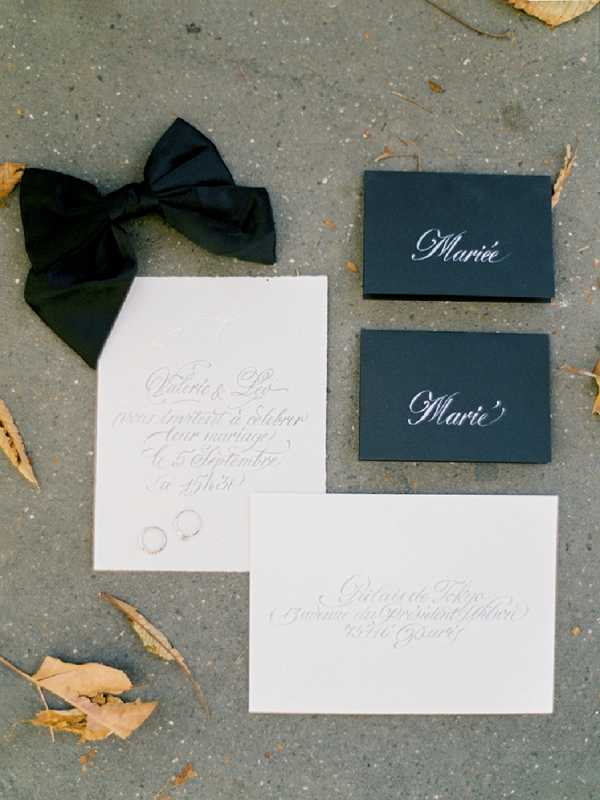 A flat lay detail shot of a French wedding stationery suite arranged on a concrete surface with scattered dried autumn leaves. The suite includes two white deckle-edge cards with grey calligraphy script — one appearing to be a wedding invitation for 'Valérie & Léo' dated '5 Septembre' and one an addressed envelope referencing 'Château de Thoiry' — along with two dark charcoal/black place cards with white calligraphy reading 'Mariée' and 'Marié.' A large black satin ribbon bow is placed to the upper left, and two delicate gold rings rest on the invitation card. The overall design palette is monochromatic black, white, and charcoal with a classic, formal calligraphy style.