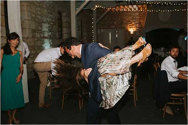 A man in a navy suit is dramatically dipping a woman wearing a floral or metallic patterned midi dress and flat sandals on a dance floor, her hair swinging downward from the motion. The scene is an indoor evening reception in a rustic stone-walled venue, likely a barn or converted outbuilding, decorated with warm fairy lights strung across the ceiling. A woman in a teal green dress watches and smiles in the background, while other guests are seated at wooden chairs in the dimly lit space. The wide shot captures the lively, informal atmosphere of the reception dancing portion of the evening.