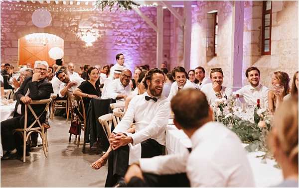 Wedding reception speeches or entertainment moment captured in a wide shot, showing approximately 30-40 guests seated at long dining tables inside a stone-walled venue with high ceilings, likely a converted barn or chateau outbuilding. The groom, wearing a white dress shirt and black bow tie, sits at the head table laughing openly, while guests around him react with smiles and laughter. The decor features warm amber uplighting on the stone walls accented with purple wash lighting, string lights along the ceiling, and table centerpieces of greenery with what appear to be blush and white florals. Guests are dressed in smart-casual to formal attire, with a mix of black and white outfits visible throughout the room. The flooring is polished concrete and wooden cross-back chairs are used throughout, giving the setting a rustic-classic feel.
