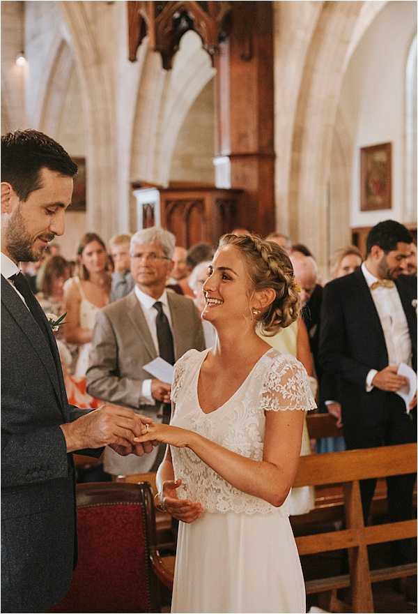 The ring exchange moment of a religious ceremony taking place inside a stone Gothic church with vaulted arches and wooden pews. The groom, wearing a dark charcoal suit with a boutonnière, places a ring on the bride's finger while she smiles broadly at him. The bride wears a white dress with a lace cap-sleeve bodice and a flowing skirt, with her hair styled in a braided updo. Approximately 10–15 guests are visible standing in the pews behind the couple, including a man in a navy suit with a yellow bow tie. The shot is a medium portrait taken from a slight angle, with the church interior softly out of focus in the background.