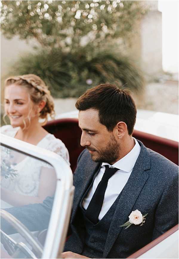 A bride and groom are seated inside a vintage convertible car with white exterior and burgundy red interior upholstery. The bride wears a lace long-sleeve dress and has her hair styled in a braided updo, and she is smiling. The groom wears a charcoal grey three-piece tweed suit with a white shirt, dark navy knit tie, and a boutonniere featuring a blush pink rose with small greenery sprigs. The shot is a close-up portrait framing both from roughly the shoulders up, with a stone building partially visible in the soft-focus background.