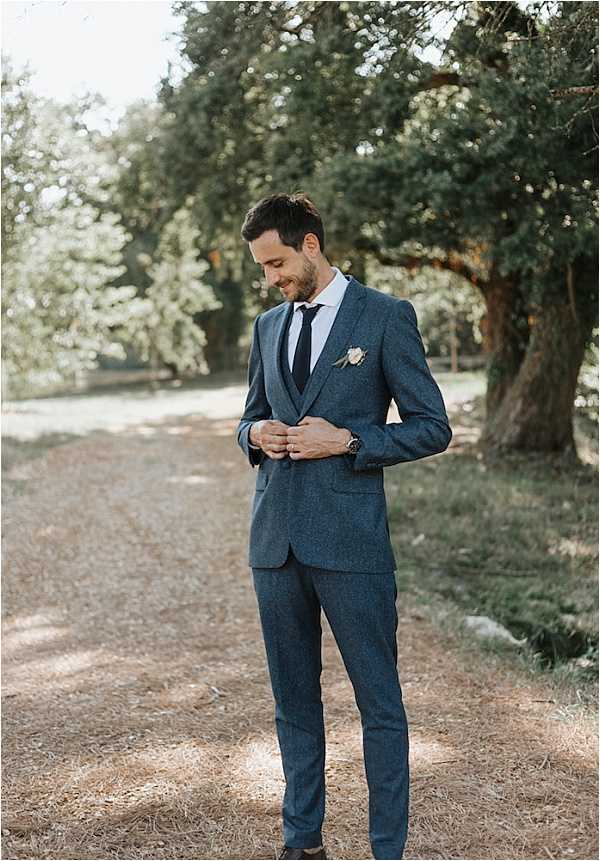A solo portrait of the groom standing outdoors on a dirt path, adjusting the buttons of his slate blue textured three-piece suit. He is wearing a navy tie, a light blue dress shirt, and a small ivory boutonniere on his lapel, along with a watch on his left wrist. The shot is a full-length portrait with the groom positioned slightly off-center, looking downward with a relaxed expression. The styling has a classic yet relaxed feel with the textured fabric of the suit adding visual interest.