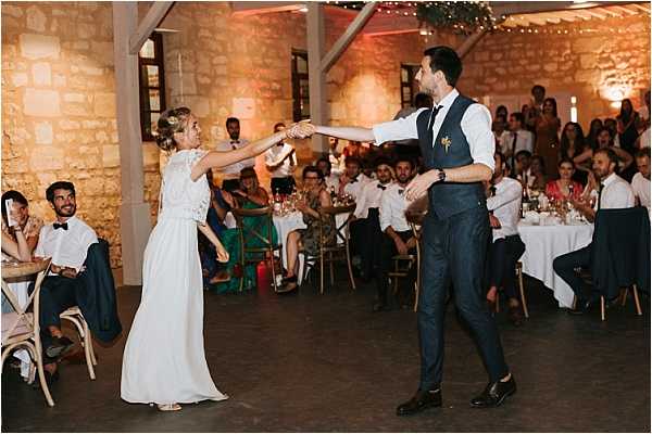 The bride and groom perform their first dance on a stone-floored dance floor inside a rustic barn or stone reception hall with exposed limestone walls lit by warm amber uplighting. The bride wears a two-piece white lace and flowing skirt ensemble with her hair up, while the groom is dressed in navy trousers and a matching navy waistcoat with a white shirt and dark tie, with a small yellow floral buttonhole. Dozens of guests are seated at round tables covered in white linens in the background, watching the couple, with string lights and greenery garlands visible along the ceiling beams. The shot is a wide, candid mid-distance photograph capturing the full dance floor and the lively atmosphere of the reception.