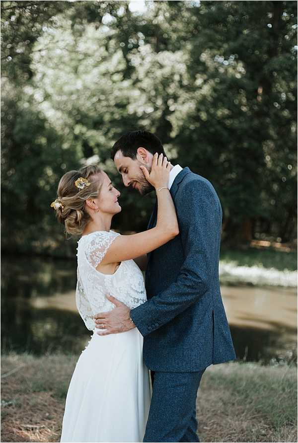 A couple portrait taken outdoors in a garden or wooded setting, with the bride and groom facing each other in an intimate pose. The bride wears a two-piece white gown with a lace cap-sleeve top and a flowing chiffon skirt, with her hair styled in a low updo accented by small gold floral hair accessories. The groom wears a fitted navy blue textured suit with a white dress shirt. The bride's hand is gently cupping the groom's face as they lean toward each other. The shot is a mid-length portrait with a shallow depth of field, leaving the tree-lined background softly blurred.