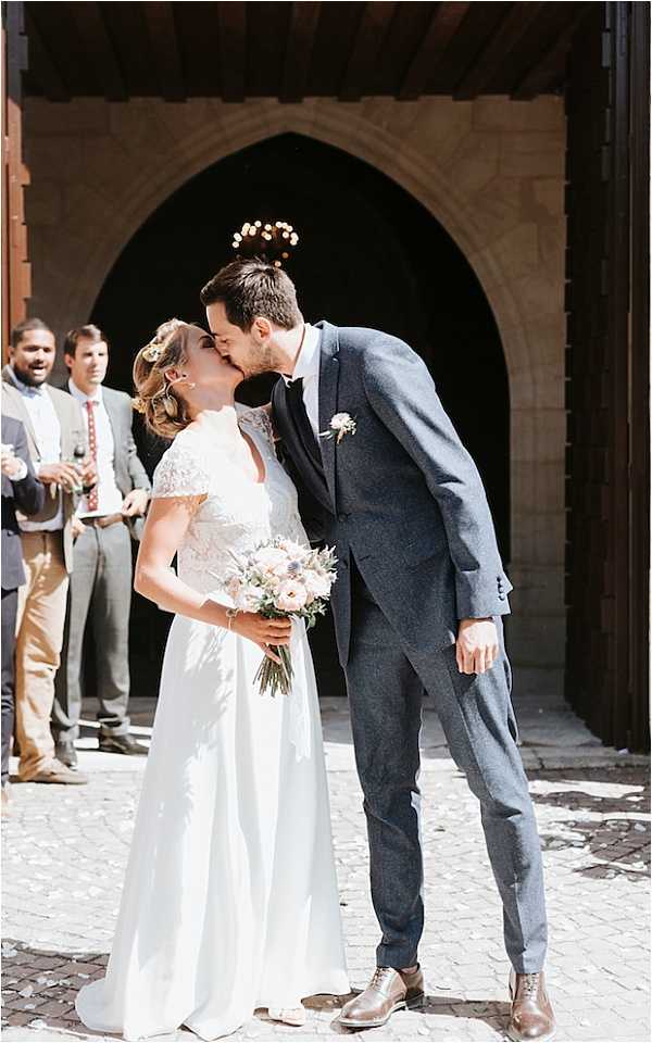 A couple shares a kiss outside the entrance of a stone chapel or church with a pointed Gothic arch doorway, likely immediately following the ceremony exit. The bride wears a white gown with a lace cap-sleeve bodice and a floral hair accessory, and holds a loose bouquet of blush ranunculus, thistles, and greenery. The groom wears a fitted navy-blue suit with a small boutonniere and brown oxford shoes. Confetti petals are scattered on the cobblestone ground around them. Several male guests in grey and tan suits are visible in the background, smiling and holding glasses. The shot is a full-length portrait taken in bright natural sunlight.