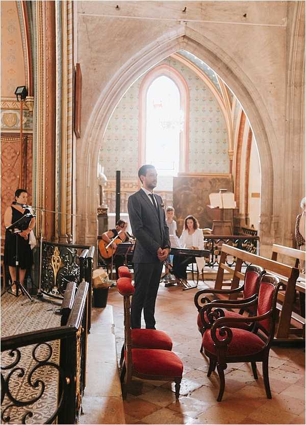 A groom stands at the altar of a historic French church during a wedding ceremony, facing forward with his hands clasped in front of him. He is wearing a dark navy suit with a tie and a small boutonniere. The church interior features Gothic pointed stone arches, decorative painted walls, ornate tile flooring, and warm natural light streaming through a tall arched window at the back. Red velvet upholstered chairs are arranged as seating in the nave, and wooden pews are visible to the right. Several musicians are set up to the left, including a guitarist, with a woman appearing to sing or officiate nearby. The composition is a medium-wide portrait shot taken from the side aisle looking toward the altar.