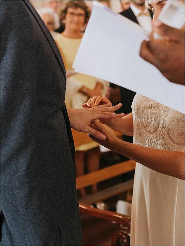 Close-up detail shot of a ring exchange during an indoor religious ceremony, likely inside a church, with wooden pews visible in the background. The bride, wearing a long-sleeved lace and crepe ivory gown, is placing a gold band onto the groom's finger; the groom is wearing a charcoal grey suit. A white ceremony booklet is partially visible in the bride's other hand. Several seated guests are softly blurred in the background, including a woman in a yellow dress.