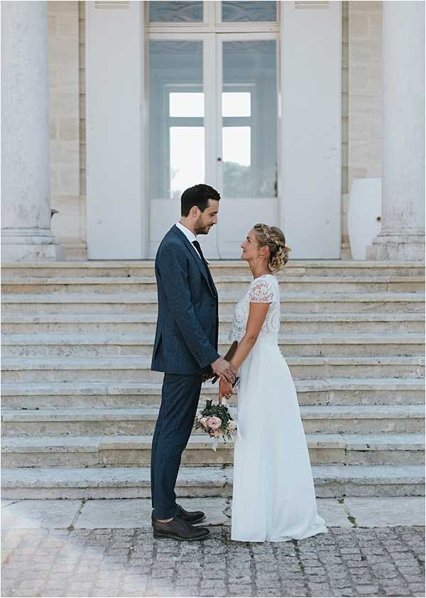 A couple portrait taken outdoors on the stone steps of a classical French venue, likely a chateau or historic building, with large white double doors and stone columns visible in the background. The bride and groom stand face to face holding hands, with the groom wearing a navy blue textured suit with a dark tie and brown oxford shoes, and the bride wearing a white gown with a lace cap-sleeve bodice and flowing skirt, her hair styled in a braided updo. The bride holds a small bouquet of blush pink flowers with greenery at her side. The composition is a medium portrait shot with a classic, understated styling approach. Potential venue feature image.