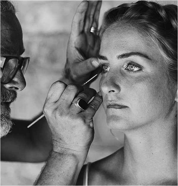 A black-and-white close-up portrait of a bride having her makeup applied by a makeup artist. The makeup artist, a bearded man wearing glasses and a large ring, is using a fine brush near the bride's eye. The bride has her hair styled in a braided updo and is looking slightly upward with a calm expression. The image is a tight, high-contrast shot with sharp detail on both subjects' faces, captured during the bridal getting-ready preparations.