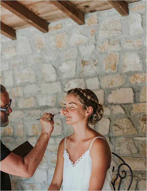 A getting-ready scene showing a bride seated in a wrought iron chair while a male makeup artist applies makeup with a brush to her face. The bride wears a white spaghetti-strap bridal slip with scalloped lace trim at the neckline, and her hair is styled in a braided updo. The setting is an indoor or covered outdoor space with exposed stone walls and rustic wooden ceiling beams, consistent with a French farmhouse or chateau property. The shot is a medium portrait framing both the bride and the makeup artist from the waist up.