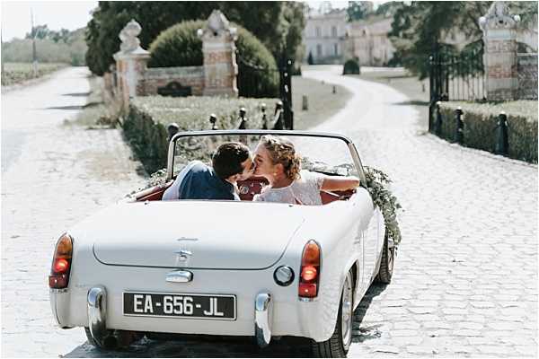 A bride and groom are kissing in the front seat of a vintage white convertible sports car, photographed from behind as it drives away along a cobblestone driveway. The car is decorated with a greenery garland along its rear. The bride wears a white lace dress and the groom a navy suit. In the background, ornate stone gate pillars and a large château building are visible at the end of the driveway, flanked by trimmed hedges and trees. Wide shot composition capturing the couple's departure. Potential venue feature image.
