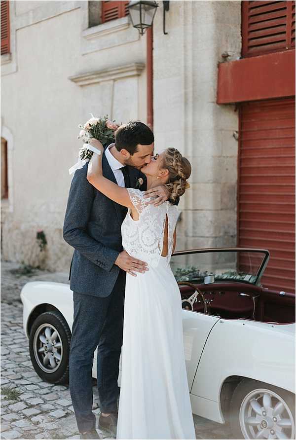 A couple portrait taken outdoors on a cobblestone street in front of a stone building with terracotta-red shutters. The bride and groom are kissing; the bride wears a white chiffon gown with an intricate lace bodice and an open-back keyhole detail, with her hair in a braided updo, and she holds a bouquet of blush and ivory blooms with greenery. The groom wears a dark navy textured suit with a white dress shirt and no tie. A vintage white convertible car with a burgundy interior is parked directly behind them, decorated with greenery along its hood. The shot is a medium full-length portrait with a relaxed, romantic feel and a rustic southern-French village aesthetic.