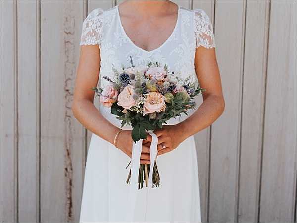 Close-up portrait of a bride from the neck down, holding a bridal bouquet in front of a weathered wooden plank backdrop. She wears a white wedding dress with a lace bodice, V-neckline, and short lace cap sleeves paired with a flowing chiffon skirt, along with a delicate gold bracelet and engagement ring. The bouquet features blush garden roses, dusty blue thistle, small mauve scabiosa, lavender sprigs, white wax flower, and mixed greenery including eucalyptus, tied with a long white ribbon streamer. The overall styling is relaxed and romantic with a soft, natural color palette.