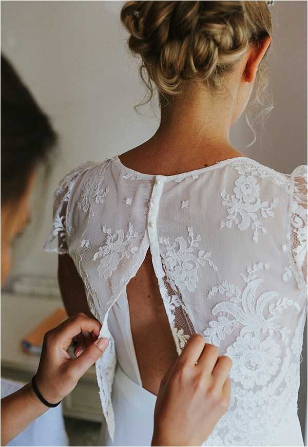 A close-up detail shot of a bride getting ready, with a second person's hands fastening the back of her white wedding dress. The gown features a sheer lace back with floral appliqué detailing, small pearl or fabric-covered buttons running down the center back, cap sleeves with lace trim, and an open keyhole or partially unfastened back panel. The bride's hair is styled in a braided updo with loose tendrils framing her neck. The shot is framed from behind at a medium-close distance, focusing on the dress construction and the dressing moment.