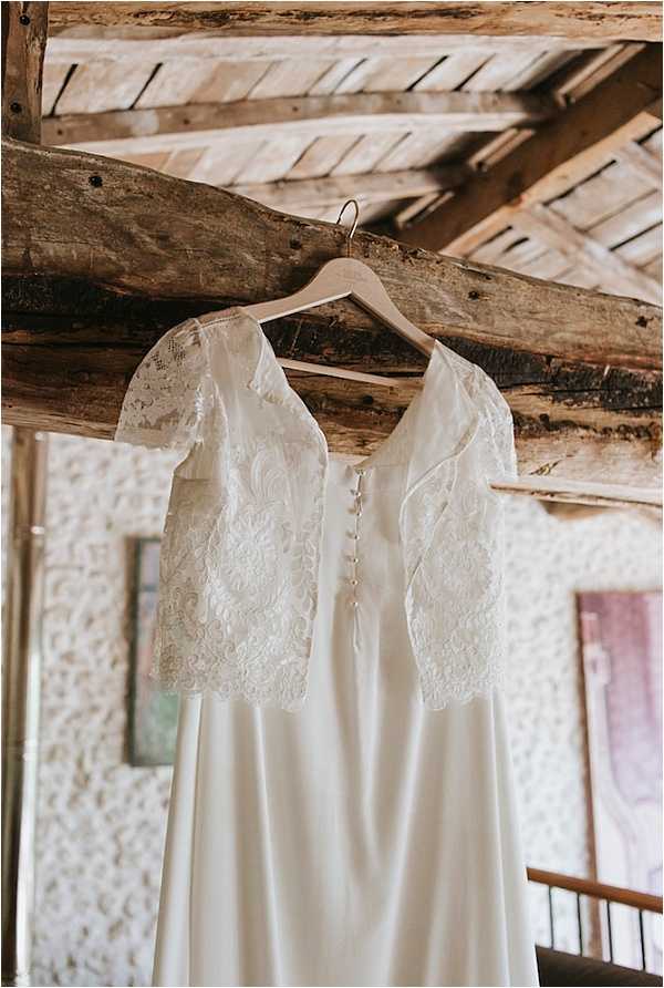 A detail close-up shot of an ivory wedding dress hanging from a wooden ceiling beam on a wooden hanger. The gown features short lace sleeves with floral embroidery, a button-back closure, and a smooth, flowing skirt. The setting appears to be a rustic interior space with exposed timber beams and textured walls visible in the soft-focus background, suggesting a barn or farmhouse-style venue.