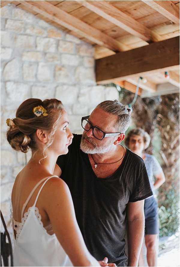 A candid getting-ready or pre-ceremony moment captured indoors beneath a wooden beam and exposed stone wall structure, suggesting a rustic venue with an open-air covered area. A bride in a white spaghetti-strap dress with lace trim detail wears her brown hair in an updo adorned with small yellow floral hair accessories. She stands facing an older man with grey-streaked hair, a beard, black-rimmed glasses, and a black t-shirt with a cross necklace, who appears to be a father figure or close family member sharing a quiet moment with her. A third person, partially visible in the background, observes the scene. The composition is a medium portrait shot with a shallow depth of field, keeping the foreground pair in focus.