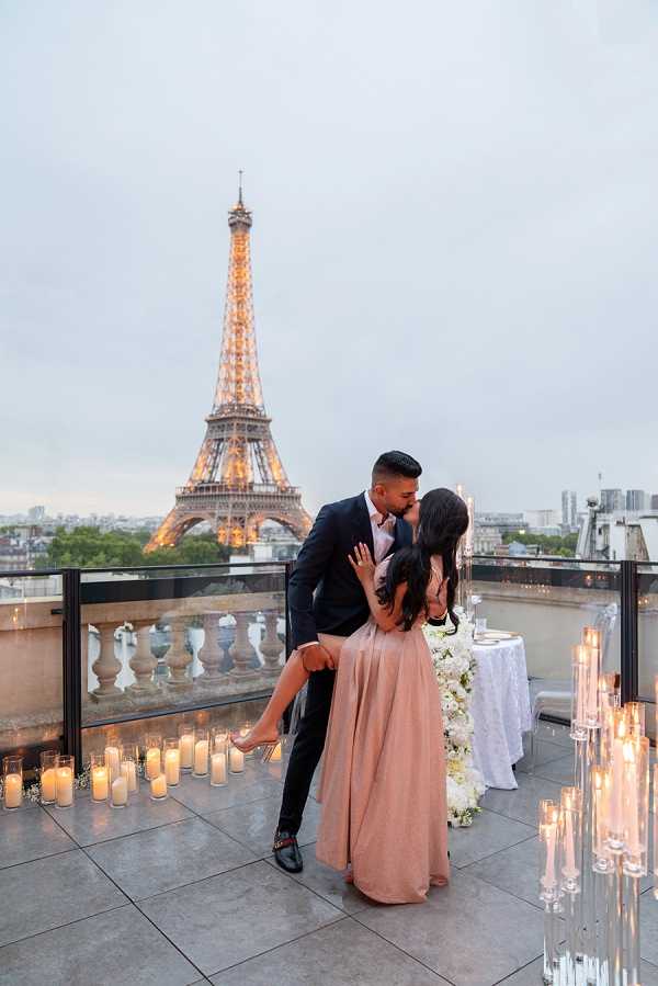 A couple shares a kiss on an outdoor rooftop terrace in Paris, with the illuminated Eiffel Tower prominently visible in the background at dusk. The woman wears a blush pink full-length skirt and the man is dressed in a dark navy suit; he dips her slightly as they embrace. The terrace is decorated with numerous pillar candles and tall glass candleholders arranged along the perimeter, alongside a white linen-covered table adorned with white floral arrangements including what appear to be white roses and hydrangeas. This is a wide portrait shot capturing both the couple and the iconic Parisian backdrop.