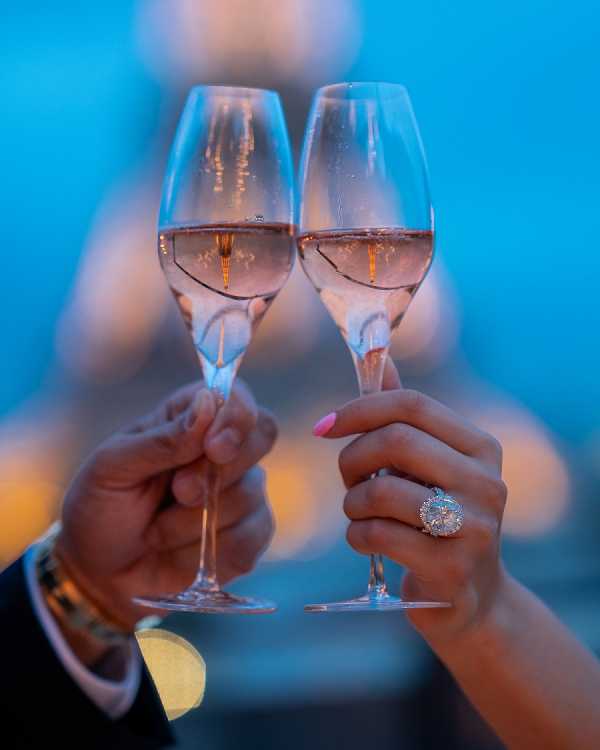 Close-up detail shot of two hands raising champagne flutes in a toast, likely during a cocktail hour or reception celebration. The bride's hand, featuring a large oval diamond engagement ring and pink manicured nails, holds one flute, while the groom's hand, partially visible in a dark suit jacket, holds the other. The flutes contain rosé champagne, and warm golden bokeh lights are visible in the blurred background against a cool blue ambient light, creating a strong contrast between warm and cool tones. The image is tightly framed on the glasses and hands, emphasizing the ring as a focal detail.