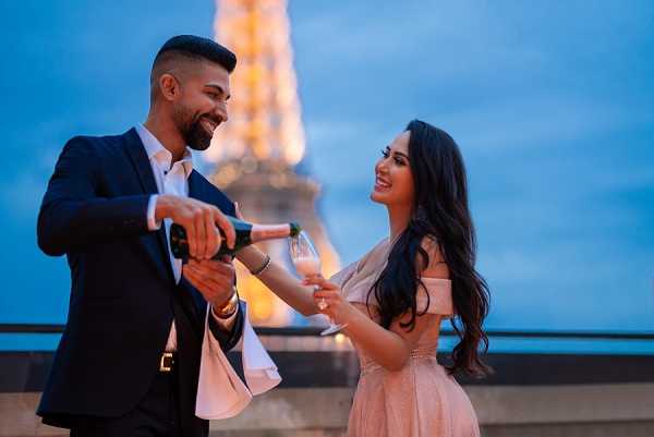 A couple poses outdoors on a terrace or rooftop in Paris at dusk, with the illuminated Eiffel Tower visible in the background against a deep blue evening sky. The man, wearing a navy suit with a white dress shirt, is pouring champagne into a glass held by the woman, who is dressed in an off-the-shoulder blush pink midi dress with her long dark hair worn down in waves. Both are smiling and facing each other in a relaxed, celebratory moment. The shot is a medium portrait framing the two figures from approximately the waist up, with the Eiffel Tower's warm golden lighting centered between them.