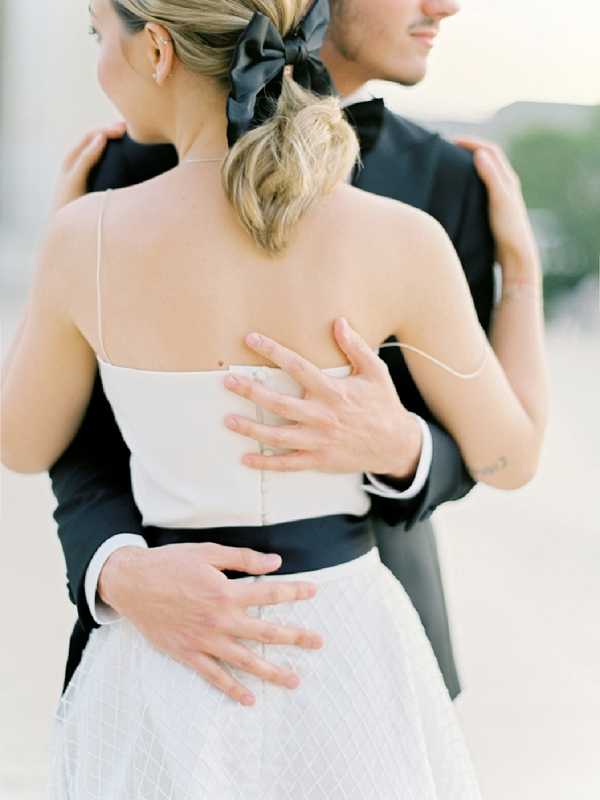 A close-up portrait of a couple embracing, shot from behind, with a soft blurred outdoor background. The bride wears a structured ivory spaghetti-strap bodice with a black waistband detail and a white textured skirt, with her blonde hair styled in a low ponytail tied with a large black satin bow. The groom wears a black tuxedo jacket with a white dress shirt, and his hands are placed on the bride's back and waist. The styling follows a modern black-and-white color palette with clean, minimal design choices.