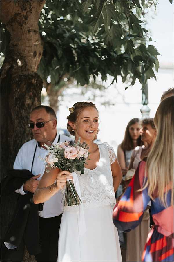 A bride is photographed outdoors, smiling and holding her bouquet while surrounded by guests, likely just before or after a ceremony. She wears a white two-piece dress with a lace cap-sleeve bodice and a flowy skirt, with small floral hair accessories and gold earrings. Her bouquet is composed of blush pink roses, white blooms, blue thistles, and eucalyptus greenery tied with a white ribbon. Several guests are visible in the background, including a man in a dark suit with sunglasses, and the setting appears to be a shaded outdoor area. The shot is a candid mid-length portrait with natural dappled sunlight.