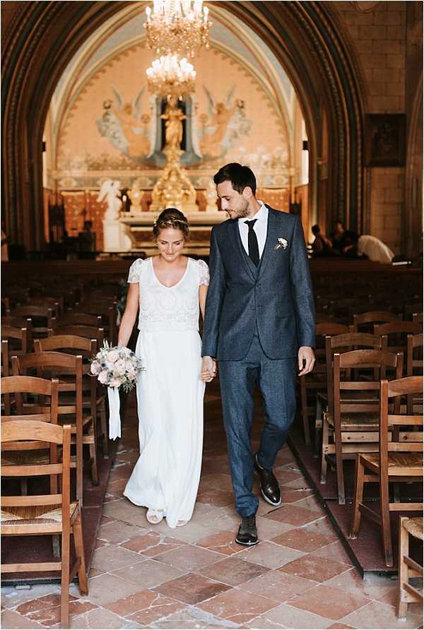 A couple walks hand-in-hand down the aisle of a French Catholic chapel following their ceremony, moving toward the camera in a medium portrait shot. The bride wears a white two-piece gown with a lace cap-sleeve top and a flowing skirt, accessorized with a delicate headband, and carries a loose bouquet of blush pink and dusty blue blooms with trailing white ribbon. The groom wears a charcoal blue three-piece suit with a black tie and a small white boutonniere. Behind them, the chapel interior features a painted apse with gold altar pieces, a crystal chandelier, and rows of wooden rush-seat chairs lining the stone tile aisle. A few guests remain seated in the background near the altar.