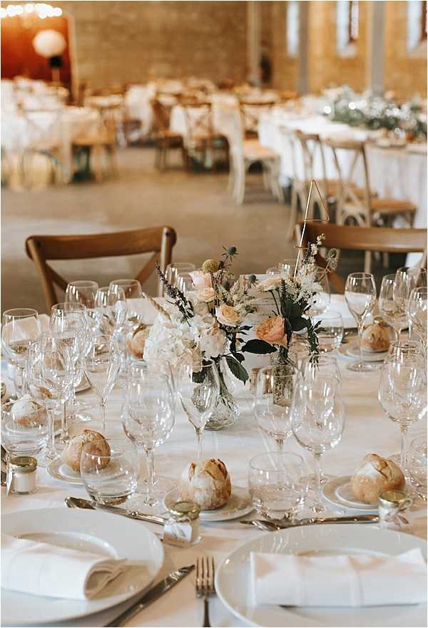 A close-up detail shot of a round reception table set for dinner inside a converted barn or stone-walled venue with exposed masonry walls visible in the background. The table is dressed with a white linen cloth and set with white square plates, white folded napkins, silver cutlery, and multiple crystal wine and water glasses per place setting, with individual bread rolls placed on side plates. The centerpiece is a low glass vase arrangement of blush garden roses, white hydrangeas, ivory wispies, and dark foliage, accented with a small geometric brass triangle table number holder. Additional round and long rectangular tables dressed in white linens with cross-back wooden chairs are visible in the soft-focus background, along with hanging globe paper lanterns, suggesting a rustic-modern styling palette of white, blush, and natural wood tones.