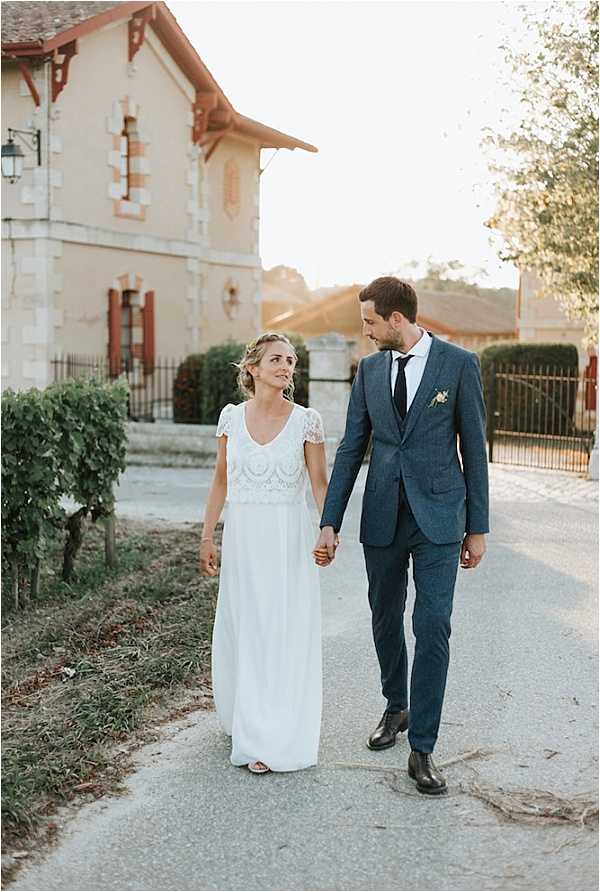 A couple portrait taken outdoors on a gravel path during golden hour, with the couple walking hand in hand and looking at each other. The bride wears a two-piece white gown with a short-sleeved lace embroidered crop top and a flowing white skirt, paired with flat sandals and an updo hairstyle. The groom wears a navy blue textured suit with a dark tie and a small white boutonniere. In the background, a cream-colored stone building with red-trimmed shutters and a decorative facade is visible, suggesting a French country estate or winery setting. The shot is a mid-length portrait with warm, natural backlighting creating a soft golden tone across the image.