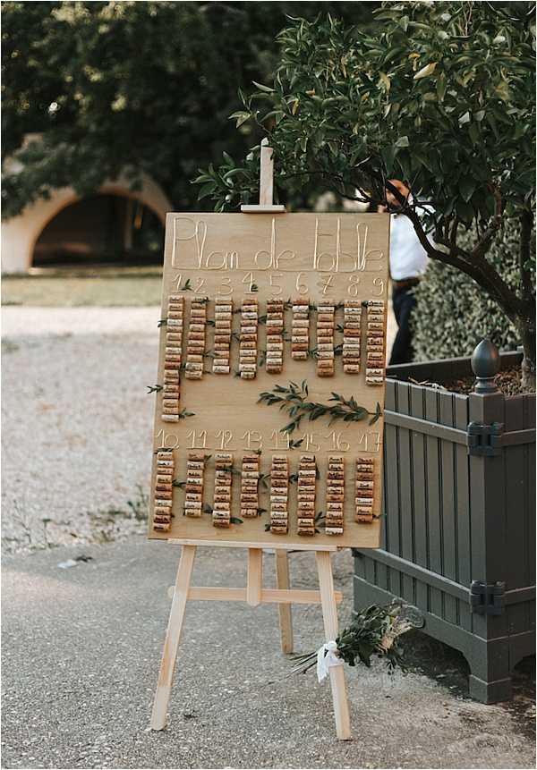 A close-up shot of an outdoor wedding seating chart ('Plan de table') displayed on a wooden easel. The board is made of light wood and features wine corks arranged in rows beneath handwritten table numbers 1 through 17, with small sprigs of olive or myrtle leaves tucked between the cork groupings as decoration. The rustic, wine-themed seating chart concept suits a French countryside aesthetic. The easel is positioned on a gravel surface beside a large dark green planter, with a blurred venue archway visible in the background.