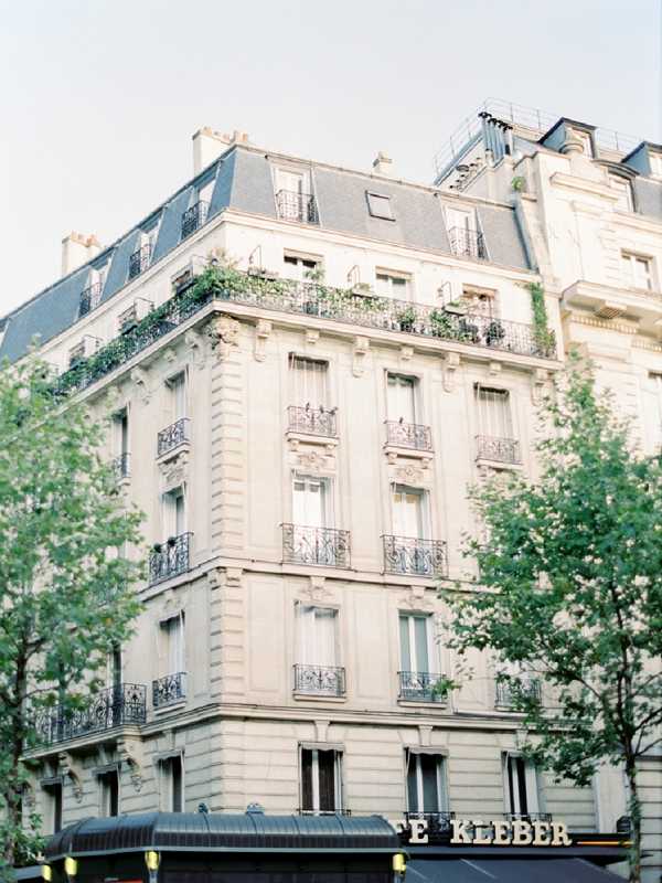 Wide exterior shot of a classic Haussmann-style Parisian building with cream stone facade, ornate wrought-iron balconies, and a mansard slate roof. The building features multiple stories of tall windows with decorative ironwork, and the upper balconies are lined with green climbing plants and window boxes. A dark awning at street level bears the text 'E KLEBER,' identifying the location. No wedding party or couple is visible in the frame. Potential venue feature image.