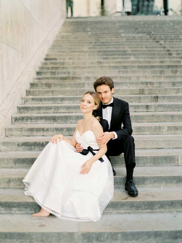 A couple portrait taken on a wide outdoor stone staircase, likely at a classical French public building or monument. The bride is seated on the steps wearing a white spaghetti-strap sweetheart ballgown with a textured skirt and a black satin sash at the waist, with her legs extended to one side; the groom sits directly behind her, wearing a black tuxedo with a black bow tie and dress shoes, his arms wrapped around her. The styling follows a classic black-and-white color palette with the contrast between the white gown and black formal wear as the central design choice. The shot is a medium-wide portrait with soft, even natural light and a film-like, slightly muted tone.