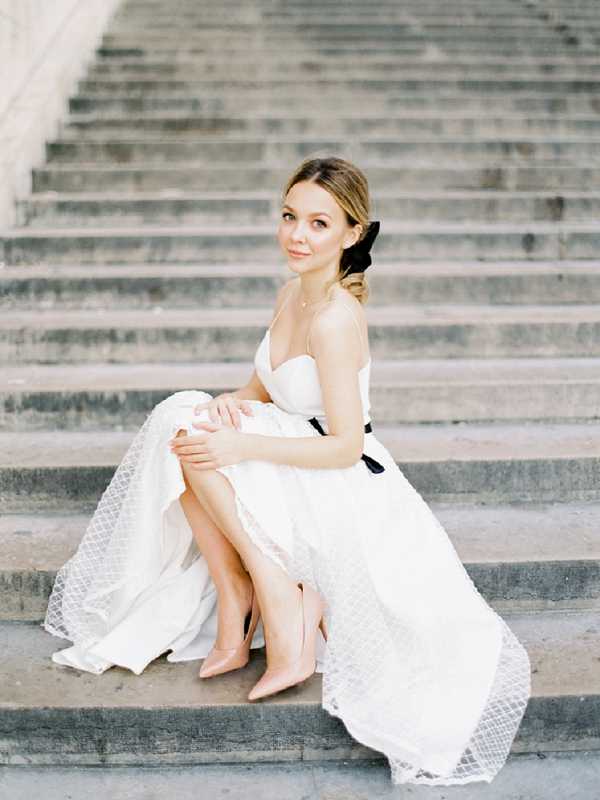 A bridal portrait of a bride seated on wide stone steps, facing the camera with a relaxed pose. She wears a white strapless ballgown with a textured lace or dotted tulle skirt and a black velvet sash belt at the waist, paired with blush pink pointed-toe heels. Her hair is styled in a low updo with a black velvet bow, and she wears a delicate necklace. The image is a medium portrait shot with soft, even natural lighting and a classic, polished styling aesthetic.