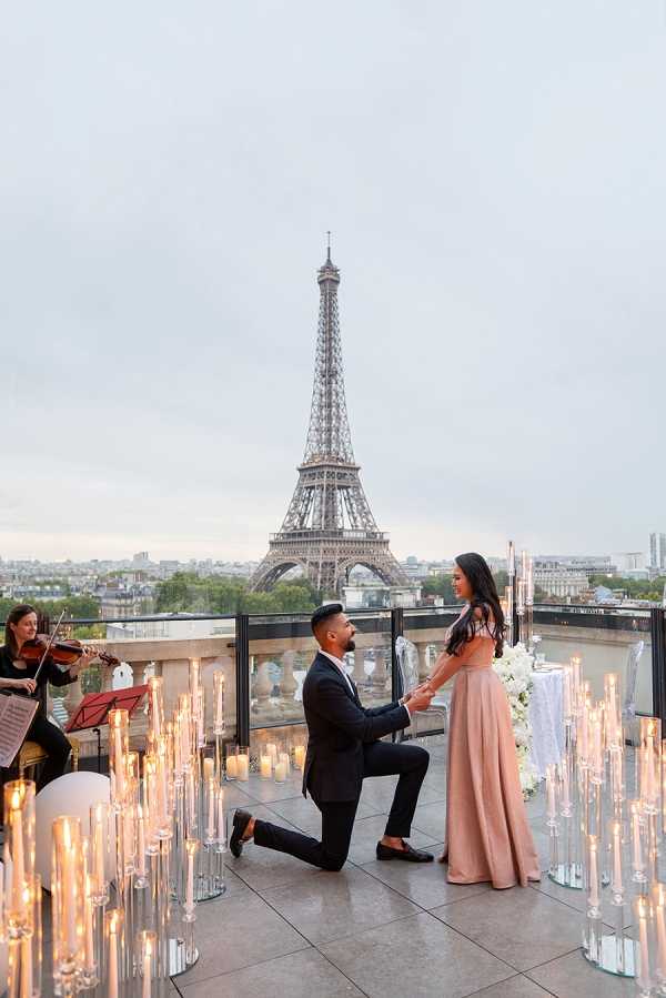 A marriage proposal taking place on an outdoor rooftop terrace in Paris, with the Eiffel Tower centered directly behind the couple. A man in a dark navy suit is down on one knee, holding the hand of a woman wearing a blush pink floor-length gown with a cutout back detail. The terrace is styled with dozens of tall glass candlestick holders and low votive candles creating a warm, glowing ambiance across the foreground. To the left, a female violinist in all black plays beside a music stand. In the background, a white-draped table with a white floral arrangement is visible. The composition is a wide portrait-style shot capturing the full scene from the couple to the iconic tower. Potential venue feature image.