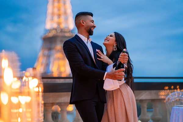 A couple celebrates on an outdoor rooftop terrace in Paris at dusk, with the illuminated Eiffel Tower visible and softly blurred in the background. The man wears a dark navy suit with a white dress shirt and no tie, while the woman wears a blush pink skirt and white long-sleeve top, with statement drop earrings and long dark hair; she is holding a champagne bottle. The two are embracing and laughing together, shot at close to medium portrait range with warm chandelier lighting visible on either side of the frame creating a golden ambient glow. The blue-hour sky and lit Eiffel Tower backdrop suggest this is an engagement or post-proposal celebration shoot rather than a wedding day itself.