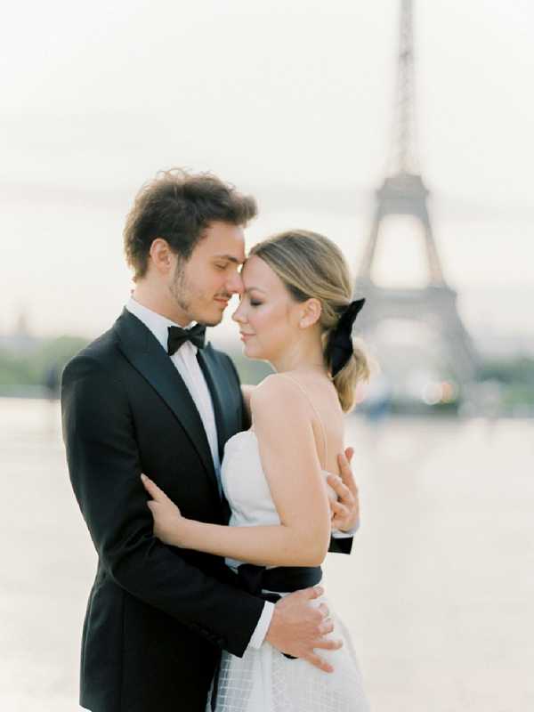 A couples portrait taken outdoors in Paris with the Eiffel Tower softly out of focus in the background. The bride and groom stand close together with foreheads nearly touching in an intimate pose; the groom wears a black tuxedo with a black bow tie, while the bride wears a white strapless gown with a black satin waist sash and has her hair pulled back with a large black bow accessory. The styling follows a classic black-and-white contrast theme with clean, minimal details. The shot is a mid-length portrait with soft, even light suggesting golden hour, giving the image a film-like, airy quality.