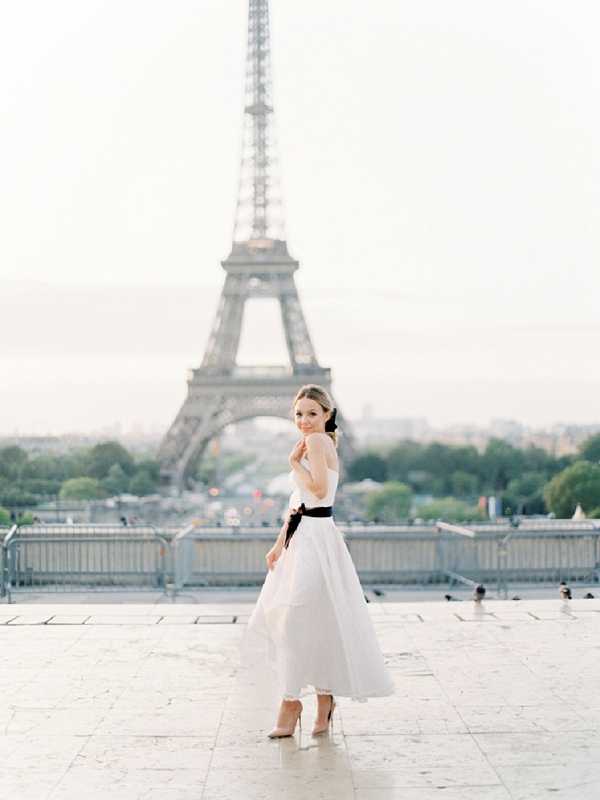 A bride poses alone on the Trocadéro esplanade in Paris, with the Eiffel Tower prominently visible in the soft-focus background. She wears a tea-length white ball gown with a structured bodice and a wide black satin sash at the waist, paired with nude pointed-toe heels and a small black hair accessory. She is turned slightly away from the camera and glances back over her shoulder, her skirt caught mid-movement. The portrait is shot at ground level with a wide depth of field that keeps the subject sharp against the hazy, overcast Parisian cityscape, giving the image a clean, classic editorial style.
