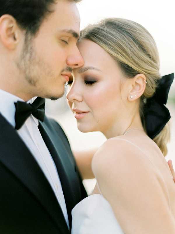 A close-up portrait of a bride and groom with their foreheads nearly touching and eyes closed in an intimate moment. The groom wears a black tuxedo with a black bow tie and white dress shirt, while the bride wears a strapless white gown and has her blonde hair styled in a low ponytail secured with a large black satin bow. She accessorizes with small pearl or diamond stud earrings and a delicate necklace. The background is completely blurred, keeping all focus on the couple's faces and the intentional black-and-white color contrast of their styling.