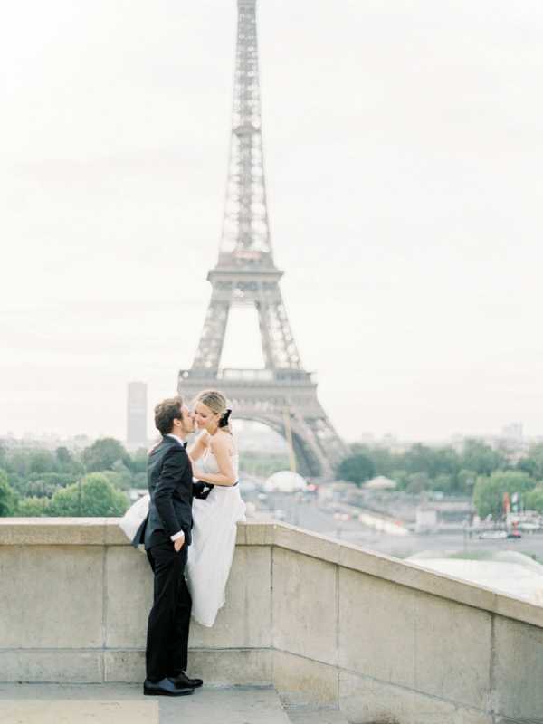 A couple portrait taken outdoors in Paris, with the Eiffel Tower prominently centered in the background. The bride, wearing a sleeveless white gown with a flowing skirt, leans against a stone balustrade while the groom, dressed in a black suit with a black tie, leans in to kiss her. The image is shot in a medium portrait composition with the couple in the lower foreground and the tower filling the background, rendered slightly soft through shallow depth of field. The overall color palette is muted and airy, consistent with film photography, giving the image a pale, low-contrast tone.