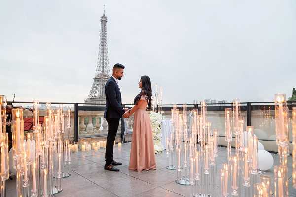 A couple stands face-to-face holding hands on a rooftop terrace in Paris, with the Eiffel Tower prominently visible in the background at dusk. The woman wears a floor-length blush pink off-the-shoulder dress and the man is dressed in a dark navy suit with black trousers. The terrace is decorated with dozens of tall glass candlestick holders at varying heights, all lit with white candles, creating a warm glowing atmosphere around the couple. A small arrangement of white florals is visible on a table in the background, and the composition is a mid-range portrait shot taken from slightly above ground level with the candle installations framing the foreground.