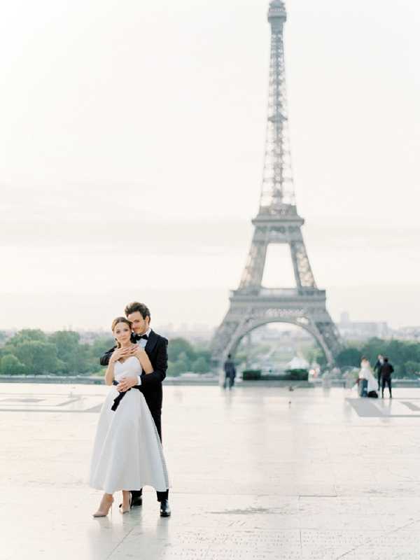 A couple portrait shot at the Trocadéro esplanade in Paris, with the Eiffel Tower centered in the soft-focus background. The groom stands behind the bride, wrapping his arms around her, both facing the camera. The bride wears a tea-length white satin ball-gown style dress with a strapless sweetheart neckline and nude heels, while the groom wears a classic black tuxedo with a bow tie. The composition is a mid-distance portrait with the couple positioned in the lower third of the frame, emphasizing the iconic Parisian backdrop. The overall styling is classic and minimalist, with a light, airy exposure giving the image a pale, film-like tone.