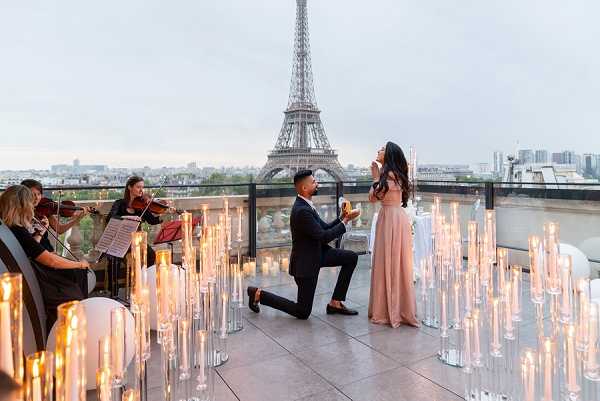 A marriage proposal taking place on a rooftop terrace in Paris, with the Eiffel Tower visible directly behind the couple in the background. The man is down on one knee proposing to a woman wearing a blush pink floor-length gown, while she reacts with her hands raised to her face. The terrace is styled with dozens of tall glass cylinder candle holders of varying heights, all lit, arranged across the floor to create a dramatic candlelit setting. Two violinists dressed in black are positioned to the left, playing for the couple during the proposal. The wide shot captures the full scene including the city skyline at dusk. Potential venue feature image.