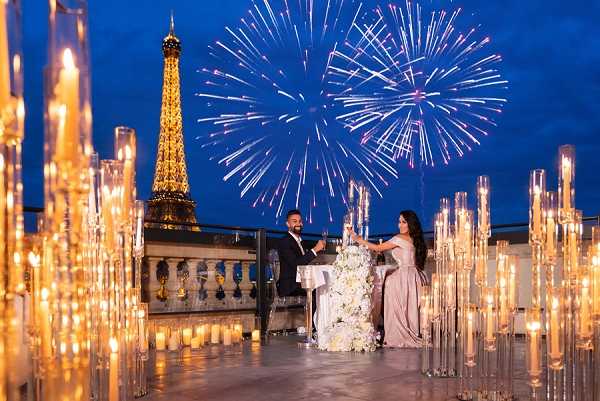 A couple shares a romantic moment on an outdoor rooftop terrace in Paris, with the illuminated Eiffel Tower and a large fireworks display visible directly behind them in the deep blue evening sky. The woman wears a floor-length dusty mauve gown and the man is dressed in a dark suit, and they stand at a small white table adorned with a tall arrangement of white flowers. The foreground is filled with rows of tall crystal candelabras and glass cylinder candle holders casting warm golden light across the terrace, creating a formal, modern decor scheme in gold and white tones. This is a wide editorial-style shot that frames the couple between the candle installations with the Parisian skyline as a dramatic backdrop.
