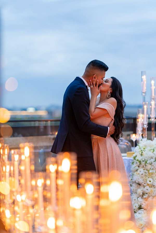 A couple shares a kiss during what appears to be an outdoor rooftop reception at dusk, with a city skyline visible in the background. The groom wears a navy suit with a bow tie, while the bride wears an off-the-shoulder blush/rose gold gown with a gold belt detail and statement drop earrings. The foreground is filled with numerous lit taper candles in tall glass holders creating a warm bokeh effect, alongside a lush arrangement of white and ivory flowers — likely roses and hydrangeas — on what appears to be a reception table. The overall decor palette is gold, blush, and white with a modern, upscale aesthetic. This is a portrait-style shot with intentional foreground bokeh from the candlelight framing the couple.