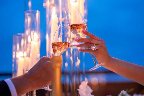 Close-up detail shot of two hands clinking glasses of rosé champagne, likely during a toast at a reception or cocktail hour. One hand belongs to a person in a white dress shirt and suit jacket, while the other hand — wearing a large diamond engagement ring and pink nail polish — belongs to the bride. The background features tall glass cylinder candle holders with lit white pillar candles and what appears to be a gold champagne bottle, creating a warm amber glow against a deep blue background. White florals are partially visible at the bottom of the frame. The overall decor palette is modern and warm, combining candlelight with a cool evening ambiance.
