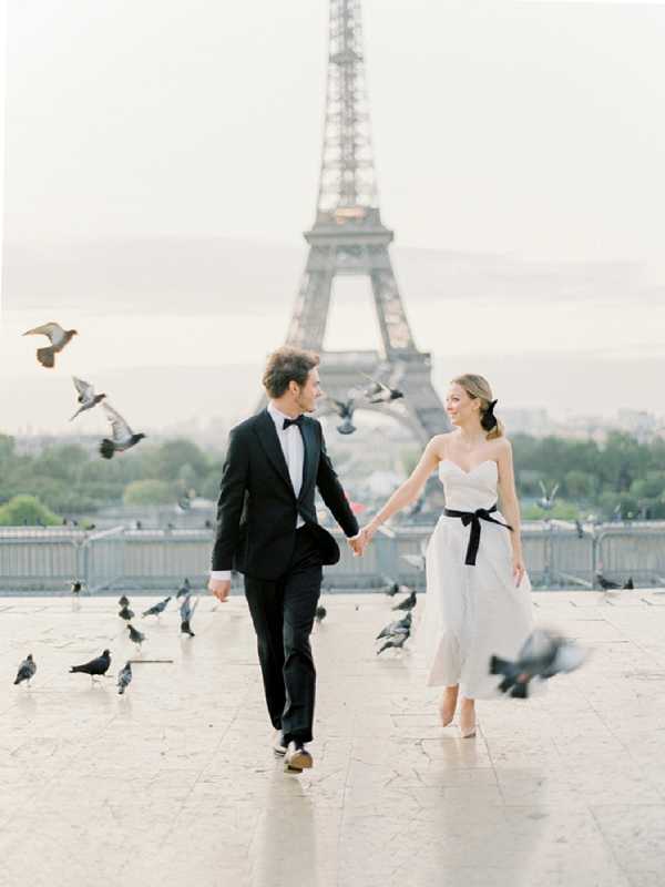 A couple walks hand-in-hand across the Trocadéro plaza in Paris, with the Eiffel Tower centered directly behind them in soft, hazy morning light. The groom wears a black tuxedo with a black bow tie, and the bride wears a strapless white midi-length dress with a bold black sash tied at the waist, paired with white heels and her hair styled in a low ponytail with a black ribbon. Pigeons are scattered and in flight around them, adding movement to the scene. The shot is a medium full-body portrait taken at ground level, with the couple slightly off-center and the Eiffel Tower serving as a strong compositional anchor.